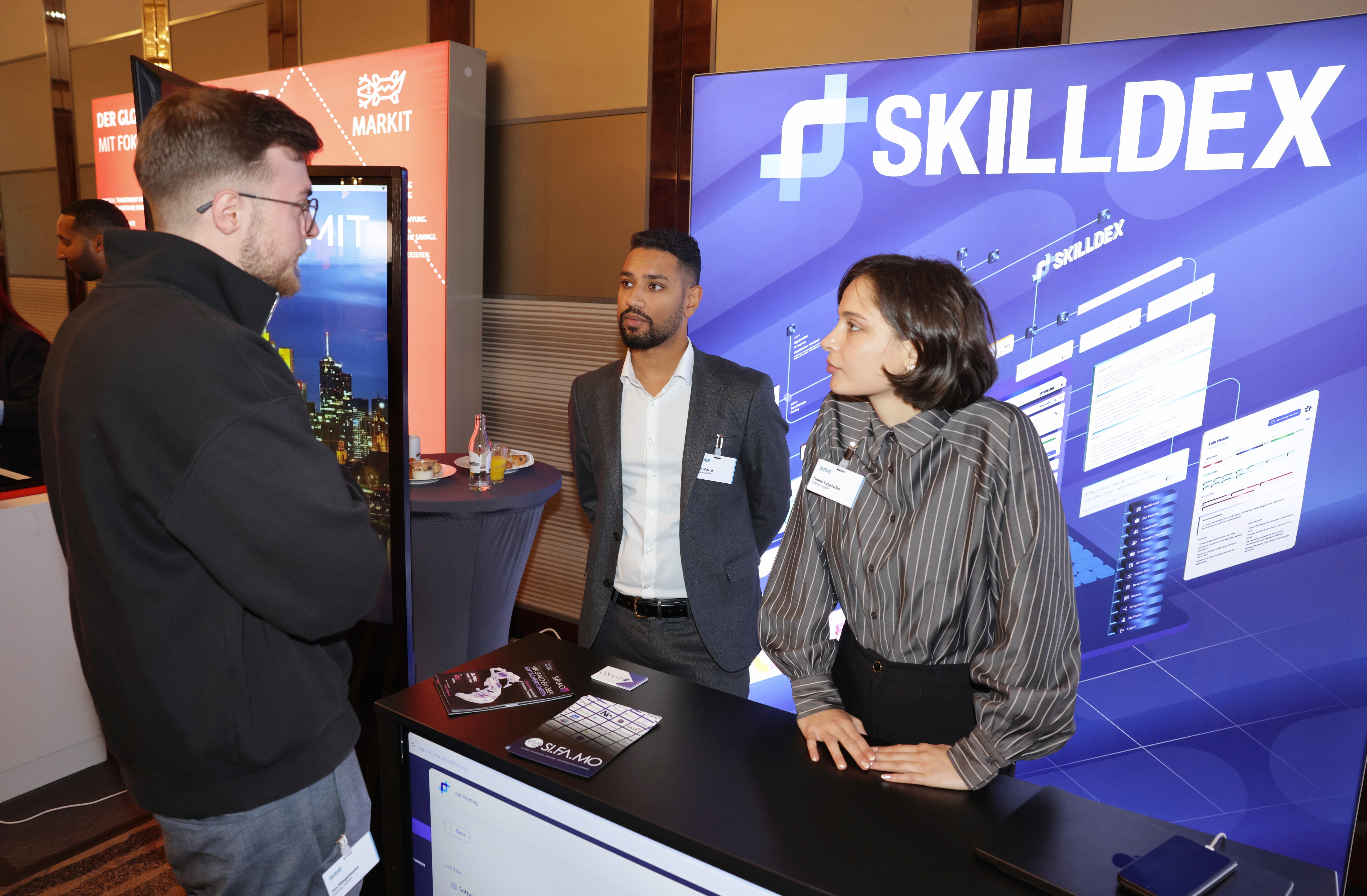 Drei Personen unterhalten sich an einem Messestand mit SKILLDEX-Hintergrund und Informationsmaterial auf dem Tisch.