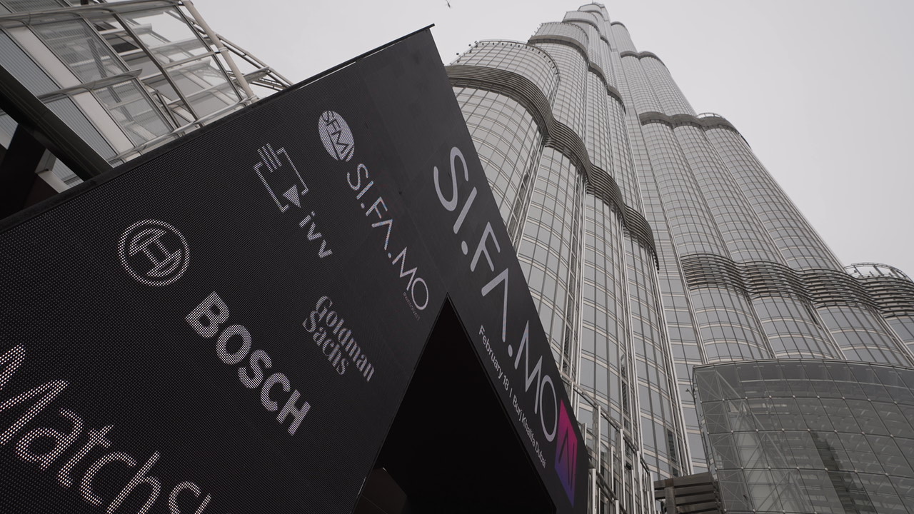 Blick nach oben auf das markante Glasgebäude Burj Khalifa mit einer großen Werbetafel für die SI.FA.MO-Konferenz und Sponsorlogos wie Bosch und Goldman Sachs im Vordergrund.