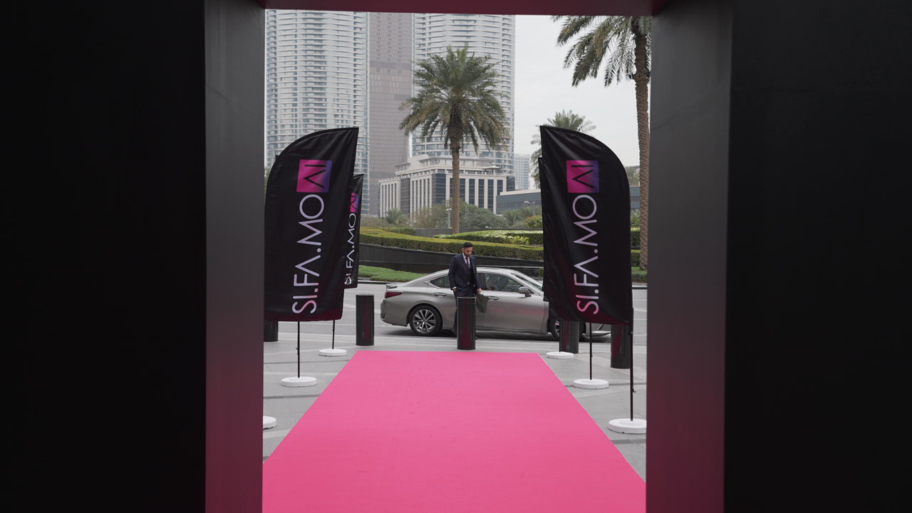 Ein Mann in einem Anzug steht neben einem silbernen Auto am Ende eines roten Teppichs mit SIFAMOW-Bannern und Palmen im Hintergrund.