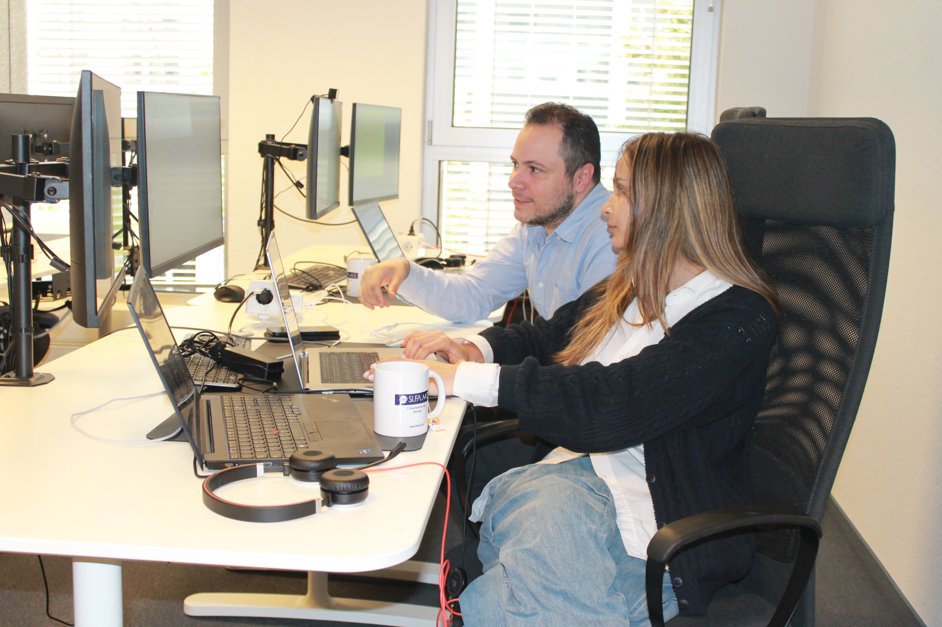 Zwei Personen sitzen nebeneinander am Schreibtisch und arbeiten gemeinsam an Laptops mit mehreren Monitoren.