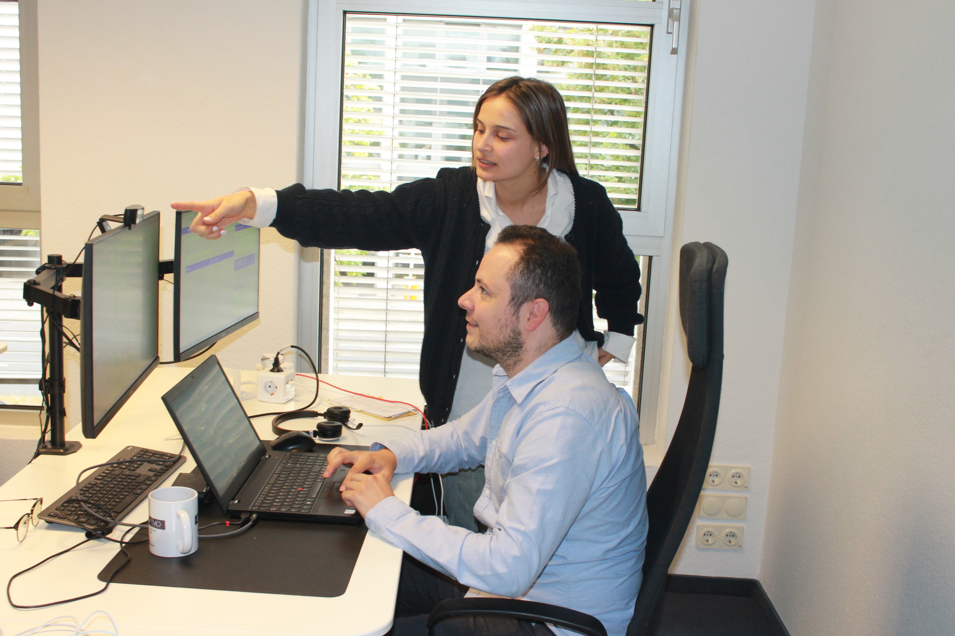Frau weist auf zwei Monitore, während ein Mann an einem Laptop in einem Büro arbeitet.