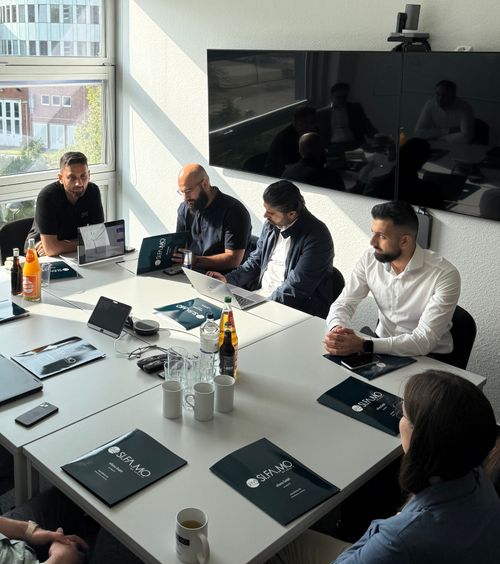 Fünf Personen sitzen um einen Besprechungstisch mit Laptops, Ordnern und Getränken in einem hellen Büro mit großem Fenster.