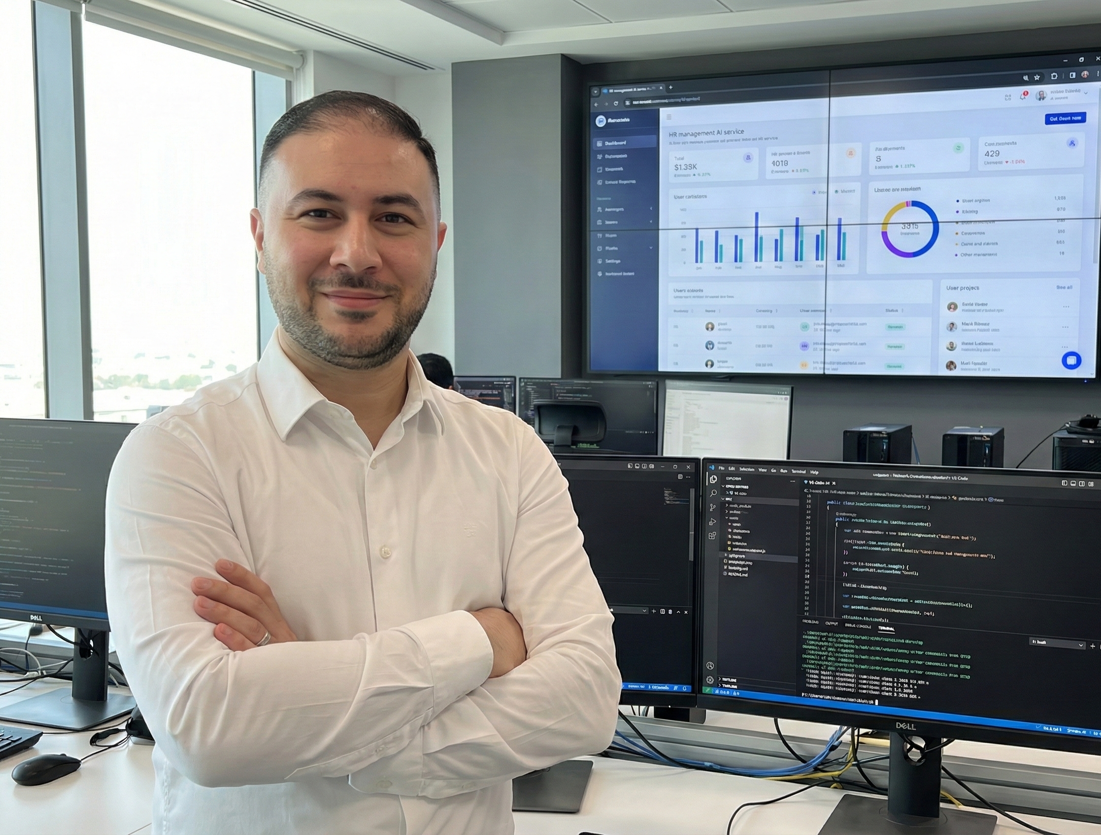 Mann mit weißem Hemd steht lächelnd vor mehreren Computermonitoren mit Codes und Diagrammen in einem Büro.