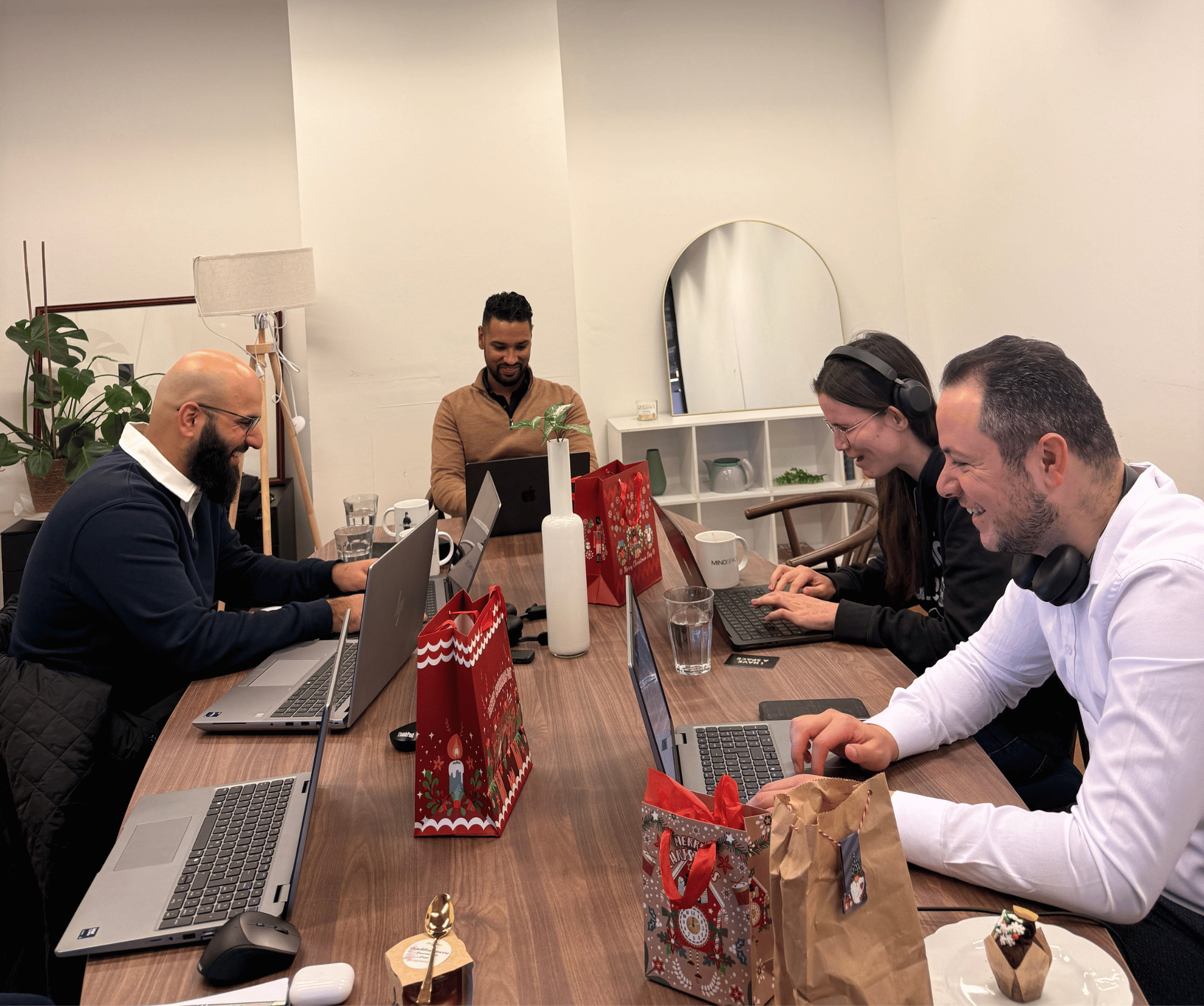 Vier Personen sitzen an einem Tisch mit Laptops, lachen und arbeiten, umgeben von Weihnachtsgeschenktüten.