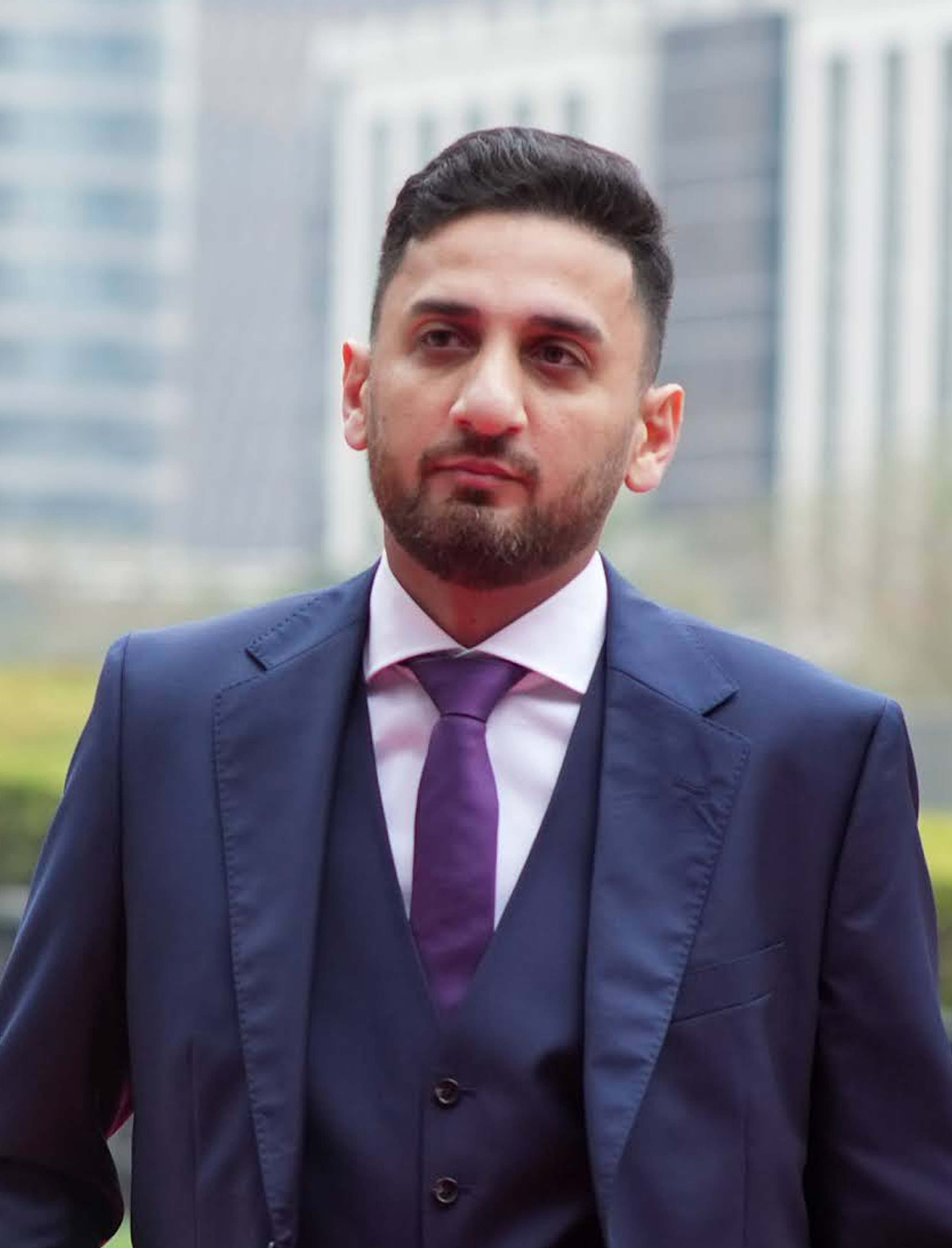 Porträt eines Mannes mit dunklem Haar und Bart in einem blauen Anzug, weißem Hemd und violetter Krawatte vor verschwommenen Gebäuden.